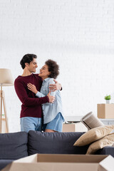 happy multiethnic couple hugging and smiling during relocation to new home