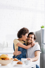Obraz na płótnie Canvas happy african american woman hugging cheerful boyfriend during breakfast