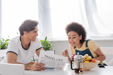 happy man holding travel life newspaper near african american girlfriend during breakfast