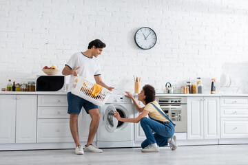 happy african american woman looking at boyfriend holding basket with dirty laundry near washing machine in kitchen