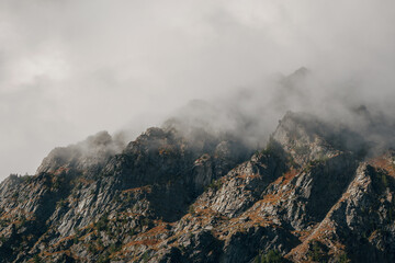 Dramatic fog among giant rocky mountains. Ghostly atmospheric view to big cliff in cloudy sky. Low clouds and beautiful rockies. Minimalist scenery mysterious place.