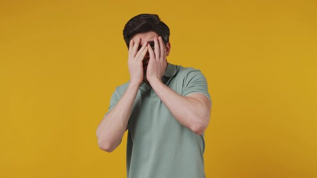 Scared shocked fun young man 20s years old wear green polo shirt look camera covering hiding face with hands peep through fingers isolated on plaine light yellow color wall background studio portrait