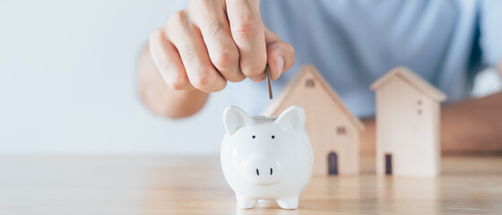 Man hand put coin into White piggy bank with wooden house on wood table. saving money for buying...