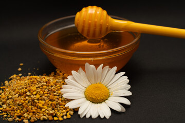 A yellow spoon of honey dipped in a jar of honey and bee bread scattered on a black background decorated with live chamomile. Honey dripping around, nice and inviting photos. Healthy food concept.