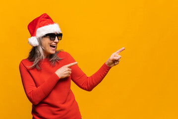 Fototapeta na wymiar Overjoyed modern mature lady wearing santa hat and sunglasses showing aside at empty copy space in ecstatic. Gray-haired senior woman recommending holiday deal point fingers away. New Year advertising