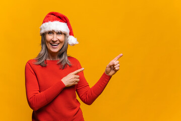 Fototapeta na wymiar Excited senior grey-haired woman wearing red santa hat points fingers aside at empty copy space, presenting novelty, advertising product. Mature lady promoting new year deal, holiday sale