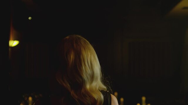 Back view of beautiful blond woman in black elegant dress  walking slowly in dark room with lighted candles . Model walking in luxury fashion show . Expensive home interior . Slow motion . 
