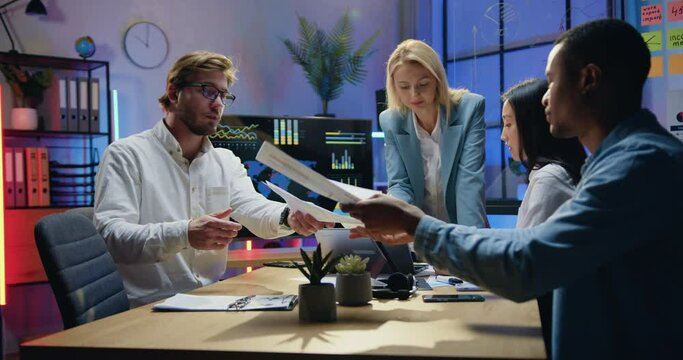 Office work concept where attractive confident smart motivated multicultural workmates browsing paper reports with charts during joint meeting in workroom in the evening