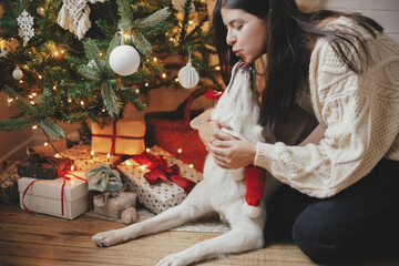 Stylish woman in cozy sweater caressing adorable dog under christmas tree with gifts and lights. Happy young female hugging and kissing cute white dog in festive scandinavian room. Happy Holidays