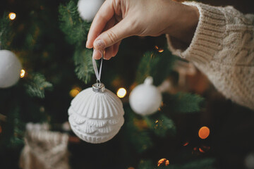 Hand in cozy sweater holding modern white bauble at christmas tree lights. Woman decorating stylish...
