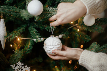 Hands in cozy sweater decorating christmas tree with modern white bauble. Woman holding vintage ornament at stylish christmas tree in lights. Winter holidays preparations. Atmospheric hygge home