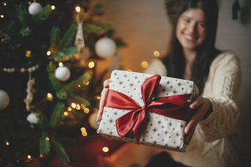 Wrapped christmas gift with red bow in happy woman hands on background of christmas tree with lights. Stylish female in cozy knitted sweater holding present in festive room. Merry Christmas