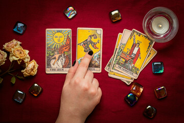 Fortuneteller's hand with black manicure lays out tarot cards, crystal, candle, dry roses on red tablecloth Flat lay Top view Fortune telling, prediction, esoteric concept - 469926211
