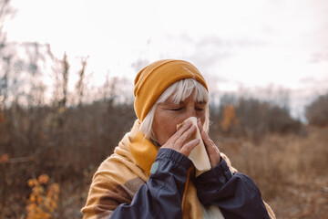 Sick middle age woman coughing and blowing her nose in park during autumn season in a cold autumn...
