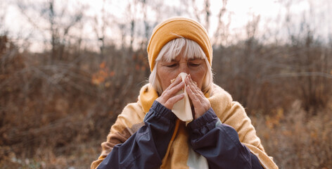 Sick adult age woman coughing and blowing her nose in park during autumn season in a cold autumn...
