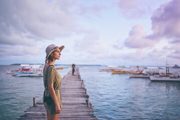 Photography and travel. Young woman in hat holding camera standing on wooden fishing pier with beautiful tropical sea view.