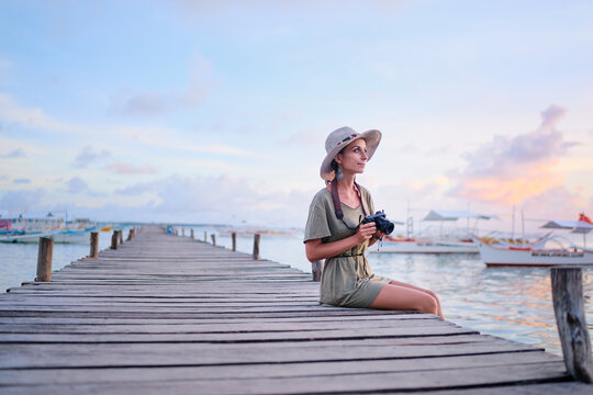 Photography and travel. Young woman in hat holding camera sitting on wooden fishing pier with beautiful tropical sea view.