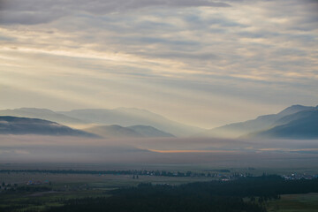 Scenic mountain landscape with orange lilac low clouds above village among mountains silhouettes under dawn cloudy sky. Atmospheric alpine scenery of countryside in low clouds in sundown golden color.