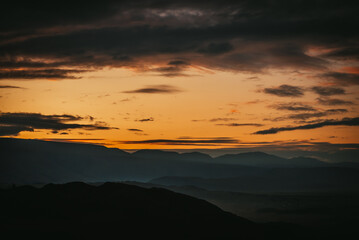 Scenic dawn mountain landscape with golden low clouds in valley among dark mountains silhouettes under sunset or sunrise sky. Vivid scenery with low clouds in mountain valley in illuminating color.