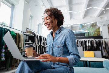 happy african american woman looking at laptop and holding paper cup in clothing boutique