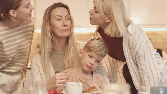 Chest-up of Caucasian woman wearing braces sitting at dining table with little son on lap, two teenage girls coming from sides and kissing mom on cheeks