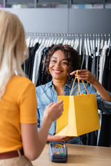 smiling african american saleswoman giving yellow paper bag with purchase to blonde client in boutique - obrazy, fototapety, plakaty