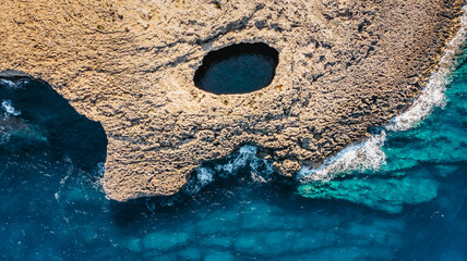Aerial view of Ahrax fallen cave,Coral Lagoon,Malta.Hole in the middle of rocks with crystal clear water inside.Rocky Maltese coast.Perfect spot for diving.Vacation in Mediterranean,travel Europe. - obrazy, fototapety, plakaty