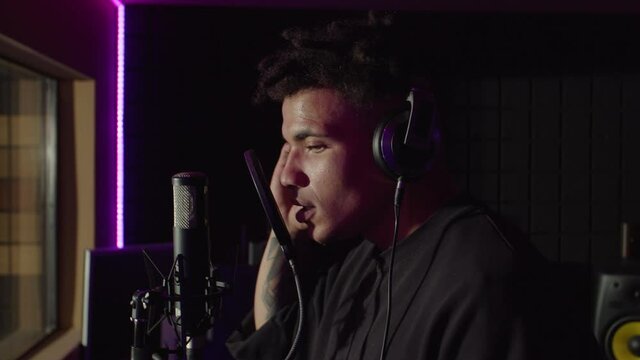 Portrait of charismatic handsome black male soul singer in headphones emotionally performing new song, singing into microphone with passion while recording hit track in music studio.