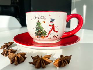 Decorated Christmas red cup of coffee with snowman next to the anise stars on the white table. Christmas drinks. Holiday morning.