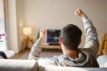Obraz premium Man on the couch is watching TV. Football fan watches football and rejoices at the victory. Entertainment and sports concept. Watching sports, movies on TV at home