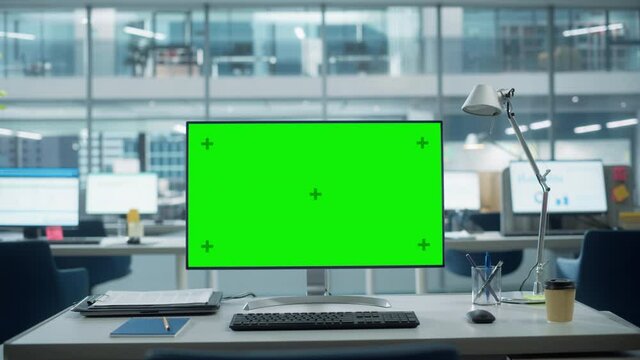 Desktop Computer Monitor with Mock Up Green Screen Chroma Key Display Standing on the Desk in the Modern Business Office. In the Background Glass Wall with Big City Office. Zoom in Shot.