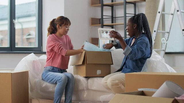 moving, people and real estate concept - women unpacking boxes and taking out book and sandglass at new home