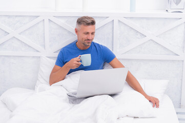 Awake from sleep. Awake man in bed. Happy guy drink tea using laptop. Morning relaxation