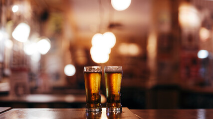 two glasses full of cold beer against the blurry empty interior of a beer bar - obrazy, fototapety, plakaty