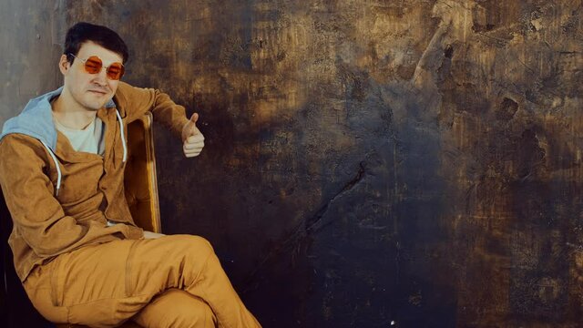 Handsome man in bright glasses sitting on chair and showing thumb up. Young male looking at camera, smiling and showing gesture of approval on background of shabby brown wall with space for text.