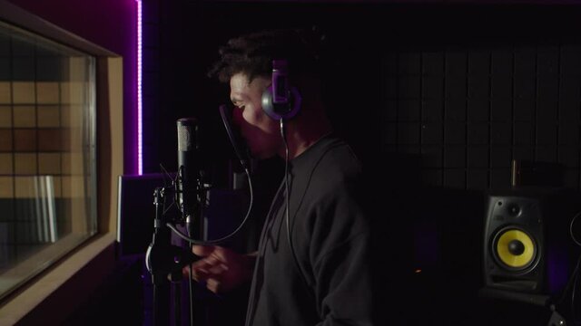 Side view of charismatic cool african american rapper with stylish haircut wearing headphones, rapping new hit song into microphone and reading lyrics on smartphone in sound recording studio.