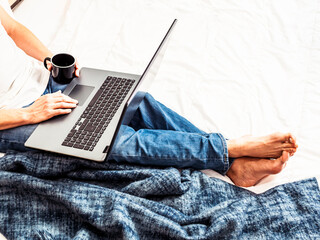 Ein Mann sitzt mit einer Tasse Kaffee und einem Laptop auf den Beinen in einem Bett. Home Office,...