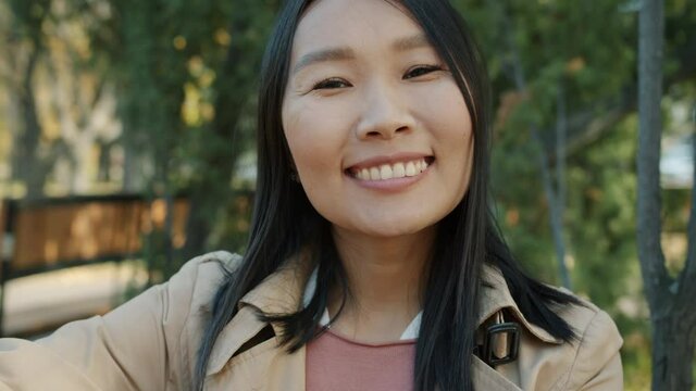 Close-up portrait of cheerful Asian girl wearing elegant clothing smiling against urban park background. Optimistic businessperson and city life concept.