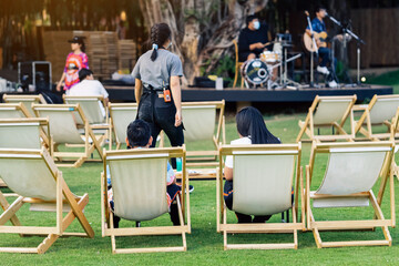 Back view of Asian young couple sit to relax on white deck chairs with tables for dinner in lawn is...