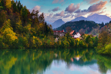 Amazing autumn landscape of a village in the Alps. View of the reflection of the fall colorful forest in the water of the river and of the mountain range in the distance at sunset.