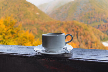 A white cup of coffee on a wooden railing against the background of an autumn landscape with sunlight. Blurry autumn background with a hot white cup. White coffee cup on autumn background.