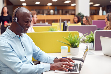 Mature Businessman Wearing Phone Headset Talking To Caller In Busy Customer Services Centre