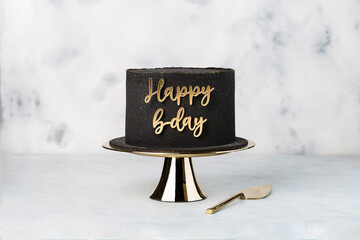 Black Birthday cake on golden cake stand. Cake on light background. topper with words 