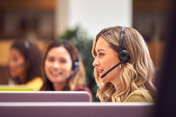 Businesswoman In Cubicle Wearing Headset Talking To Caller In Busy Customer Services Centre