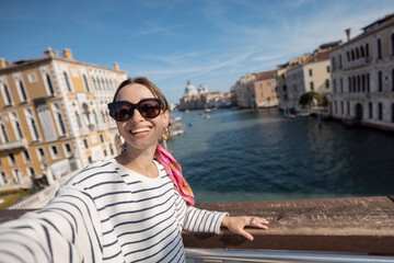 Fototapeta na wymiar Young woman taking selfie photo on the background of famous Grand Canal in Venice. Idea of spending summer time and happy travel in Italy. Caucasian female in white sunglasses and striped vest