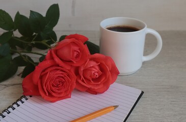 A bouquet of red roses, an empty notebook, and a mug of coffee on the table.	