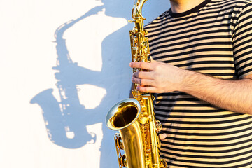 Young man in sunny outdoor place playing saxophone in front of a wall.