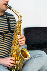 Musician sitting and playing saxophone in front of a white wall.