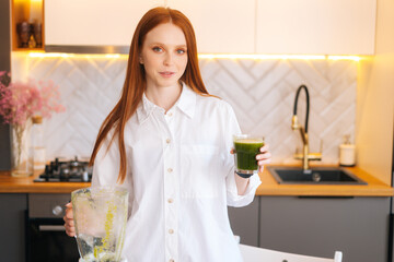 Portrait of pretty redhead young woman holding glass with green vegetable detox smoothie cocktail from blender, in kitchen with light modern interior. Concept of healthy eating and lifestyle.