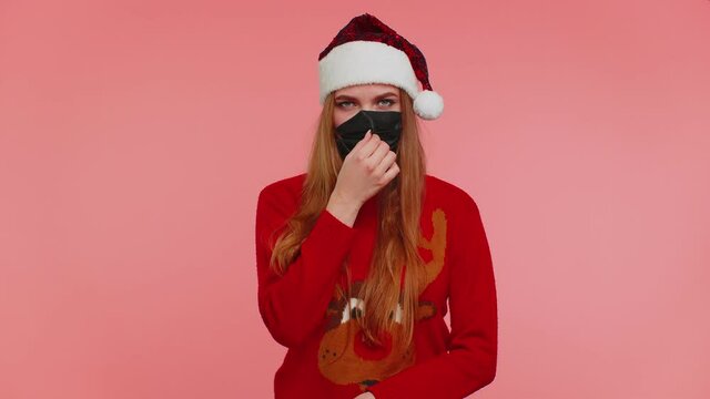 Woman in Christmas red sweater and hat wearing sterile face mask ppe to safe from coronavirus on lockdown quarantine. Adult girl indoors isolated on pink background. Covid-19 New Year celebration fail